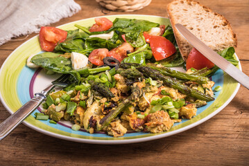 Fried scrambled eggs with asparagus, spinach and tomatoes in a large plate on a rustic wooden table, top view, close-up - simple rustic breakfast