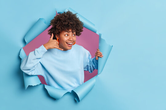 Happy cheerful Afro American woman makes call me gestue looks with interest away smiles broadly dressed in casual jumper poses in ripped paper hole of blue background blank space for your text