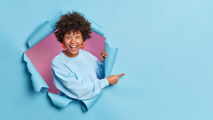 Cheerful surprised dark skinned woman breaks through paper wall demonstrates copy space aganst blue...