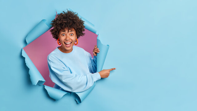 Cheerful surprised dark skinned woman breaks through paper wall demonstrates copy space aganst blue background gives advice shows advertisement on empty space wears casual sweater and earrings