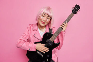 Pink haired pleased woman tilts head plays acoustic guitar enjoys favorite melody prepares for rock n roll concert wears fashionable clothes poses in studio against rosy background. Music concept