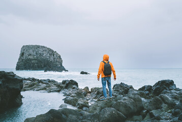 An active backpacker man dressed orange waterproof jacket with backpack enjoying Atlantic ocean bay view on rocky Madeira island seashore. Portugal.