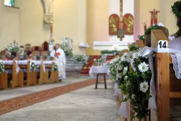 Fototapeta na wymiar Interior of church decorated for First Holy Communion celebration, benches with white flowers, priest in soft focus in front of main altar
