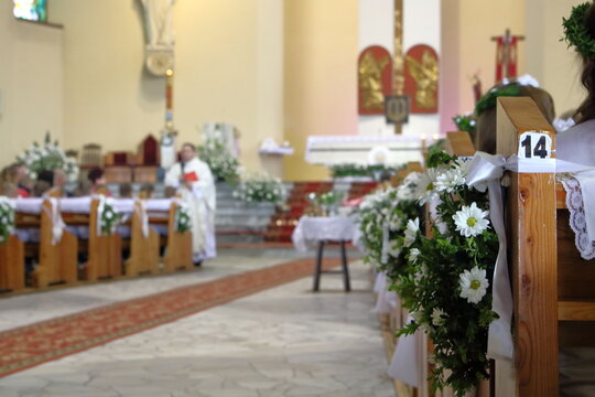Interior of church decorated for First Holy Communion celebration, benches with white flowers, priest in soft focus in front of main altar