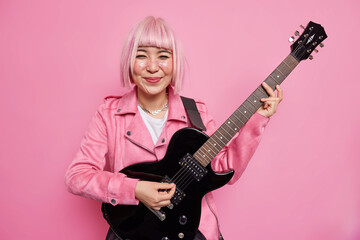 Fototapeta premium Young cheerful woman guiratist with pink bobbed hairstyle plays favorite music on acoustic electric guitar ejoys hobby and leisure activities poses indoor wears jacket. Talented musician or soloist