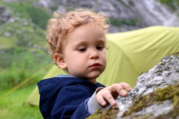 Travel with children. Cute curly two-year-old boy in front of a tent in the mountains..