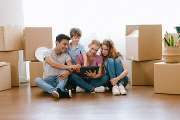 Fototapeta na wymiar Happy caucasian family with teenage daughter and younger brother sitting on floor among cardboard boxes have fun using modern tablet buying furniture into new house.E-commerce retail services concept.