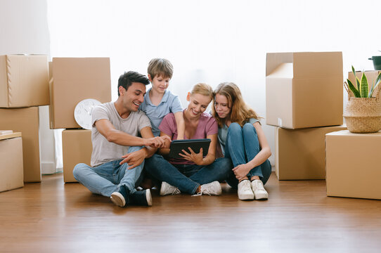 Happy Caucasian Family With Teenage Daughter And Younger Brother Sitting On Floor Among Cardboard Boxes Have Fun Using Modern Tablet Buying Furniture Into New House.E-commerce Retail Services Concept.