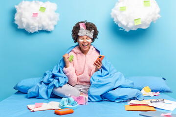 Emotional woman being on self isolation works distantly stays in bed wears slumber suit and sleepmask uses smartphone for online communication works on papers gad to finish task. Domestic atmosphere © wayhome.studio