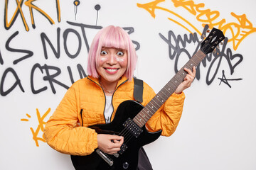 Happy surprised Asian hipster girl with trendy pink hairstyle wears orange jacket poses against tagged wall plays favorite music feels free and independent. Youth hobby and urban style concept
