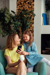 Happy caucasian mom is sitting in a chair with her daughter and watching phone