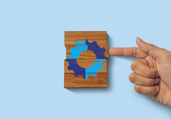 Hand putting the last piece of wooden blocks with the gear icon on blue background. Team work,...