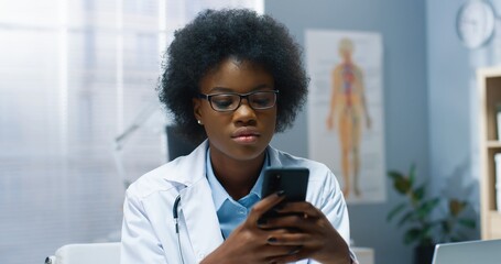 Close up portrait of African American young beautiful female doctor in glasses sitting in hospital texting on smartphone browsing online at workplace. Woman physician typing on cellphone in cabinet