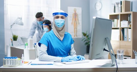 Portrait of beautiful young Arabic female healthcare specialist wearing medical mask and face shield in hospital sitting at desk in cabinet looking at camera. Woman doctor work covid pandemic