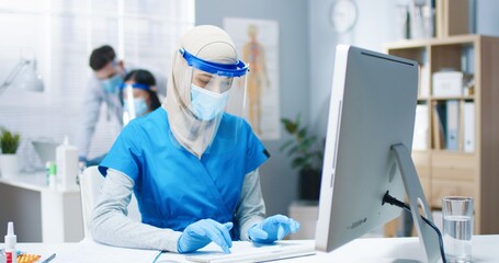 Portrait of beautiful young busy Arab female healthcare worker nurse in medical mask and face shield sitting at desk in cabinet typing on computer working in hospital. Woman doctor, covid pandemic