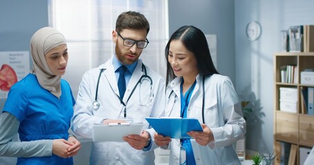 Portrait of joyful mixed-race young professional male and female doctors standing in hospital and speaking. Man physician holding paper clipboard with treatment results talking to women colleagues