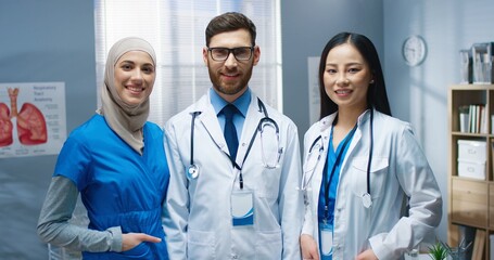 Portrait of multi-ethnic cheerful positive young doctors standing in hospital looking at camera and smiling. Team of healthcare specialists at work in clinic with smile on face. Medical center concept