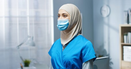 Fototapeta premium Close up of Arabic young beautiful woman general practitioner in medical mask and uniform standing in hospital looking at camera. Female pretty nurse working during covid-19 pandemic