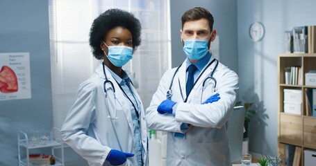 Portrait of young mixed-race male and female doctors in medical masks at work in medical center looking at camera Team of Caucasian man and African American woman healthcare workers during coronavirus
