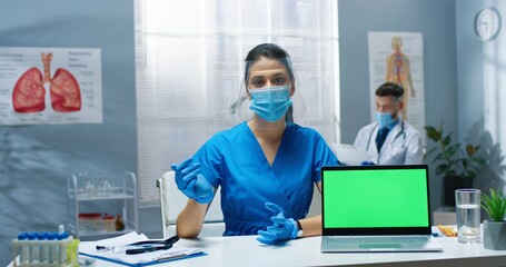Portrait of young Caucasian beautiful nurse woman in protective face shield and medical mask sitting at table in cabinet with laptop with green screen, speaking, looking at camera at consultation