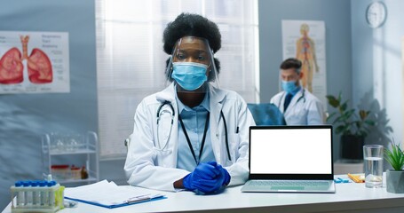 Portrait of African American beautiful female therapist in cabinet in mask and face shield sitting at desk with laptop with chroma key talking on online video consultation explaining to patient, covid