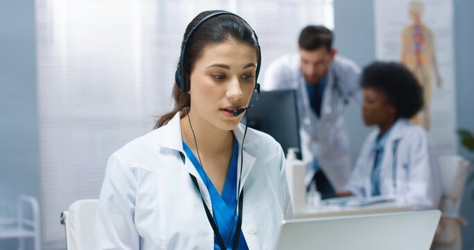 Close up of Caucasian young beautiful female doctor having online video call consultation on laptop speaking in headset with patient sitting at workplace in hospital cabinet. Healthcare, online chat