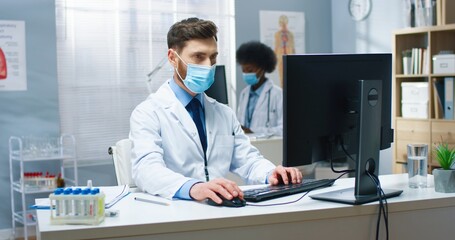 Naklejka na ściany i meble Portrait of handsome young Caucasian professional male doctor in medical mask and coat texting on computer surfing online while sitting at desk at workplace. Healthcare work, medic concept, covid-19