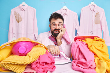 Bearded European man bites finger nails looks nervously at camera does housekeeping duties irons...