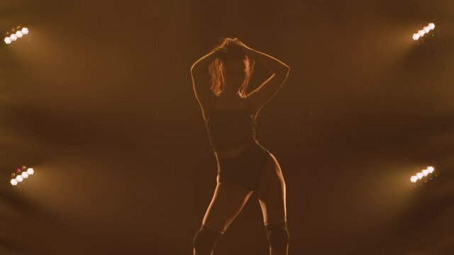 Silhouette of a sporty slender woman in black underwear dancing twerk in a dark studio with bright lights. The female dancer shakes her buttocks and seductively moves her half naked body. Slow motion.