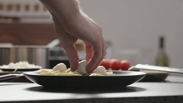 Slow motion orbit shot of man add mozzarella balls to pesto penne on black plate