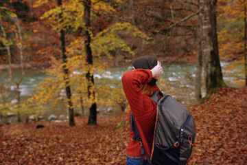 joyful woman in a hat sweater with a backpack on her back gestures with her hands in a park in nature