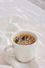 Taza blanca de porcelana labrada con café con flores blancas del olivo sobre tejido blanco en un fondo luminoso.