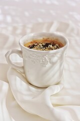 Taza blanca labrada con café con flores del olivo sobre tejido blanco en un fondo blanco y con luz.