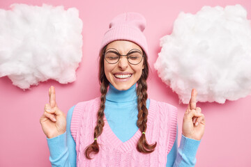 Cute happy teenage girl hopes for good luck crosses fingers anticipates positive news stands over white clouds closes eyes wears round spectacles hat and turtleneck. May my dreams come true.