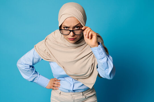 Portrait of a Muslim woman in hijab holding eyeglasses by the temple and looking carefully through them with raised eyebrow