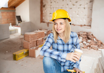 pretty young worker woman with yellow safety helmet takes coffee break on construction site indoor and is happy