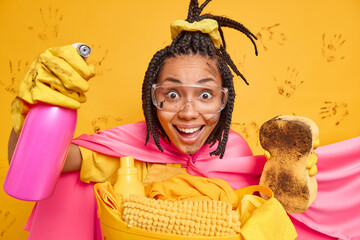 Positive ethnic woman dressed in superhero costume holds cleaning detergent and dirty sponge smiles...