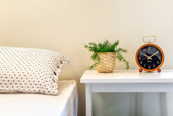 Bedside table with greenplant in straw pot and clock on it. Part of bed is visible. Interior...