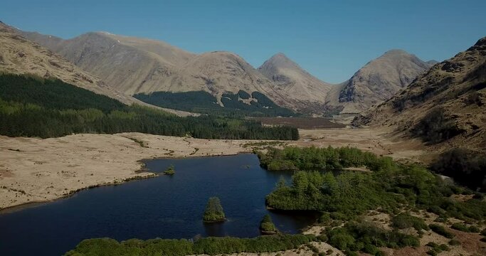 4k aerial footage of a tranquil loch in Glen Etive, Scottish Highlands with mountains and cloudless blue sky backdrop. Small island of trees in loch. No people.
