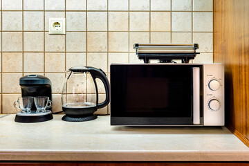 Kitchen equipment: coffee maker, boiler, microwave owen and toster in bleck colour. Tiled wall at the background. Interior element.