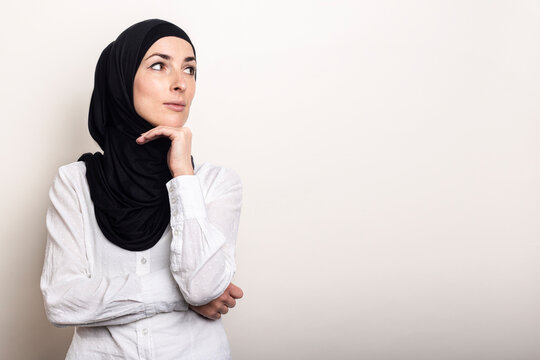 Young Muslim woman dressed in a white shirt and hijab holds her hand to her chin and looks to the side on a light background. Banner