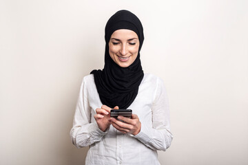 Muslim Young woman in hijab holds a phone in her hands and smiles on a light background. Banner. Writes messages