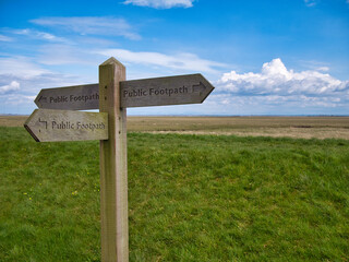 A wooden sign showing the directions of three public footpaths. Taken in a coastal setting on a sunny day with blue skies and white clouds.