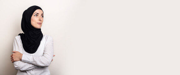 Young Muslim woman dressed in a white shirt and hijab looks to the side on a light background. Banner