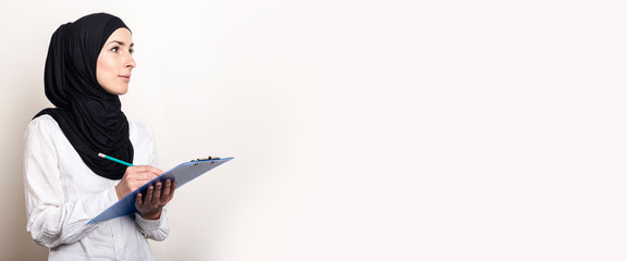 Young Muslim woman in a white shirt and hijab holds a clipboard and looks to the side. Auditor hiring. Banner