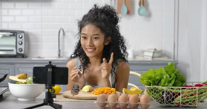 A young, happy and beautiful Asian woman with curly long hair wearing grey gingham apron. She's makes video lesson about making meal or live streaming online to show cooking method in modern kitchen.