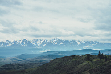 Scenic mountain landscape with trees on hill and great snowy mountain range among low clouds and forest in valley at early morning. Atmospheric alpine scenery with high mountain ridge under cloudy sky