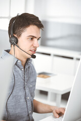 Casual dressed young man using headset and computer while talking with customers online. Call center, business concept