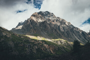 Mountain landscape with rocks with snow in sunlight and low clouds on top. Awesome rocky wall with sharp rocks in sunshine. Atmospheric mountain scenery with high rocky mountain pinnacle in clouds.