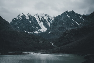 Dark atmospheric landscape with mountain lake among deep black rocks on background of snowy mountains under gray cloudy sky. Bleak view to mountain lake and glacier in deep black colors. Dark tones.